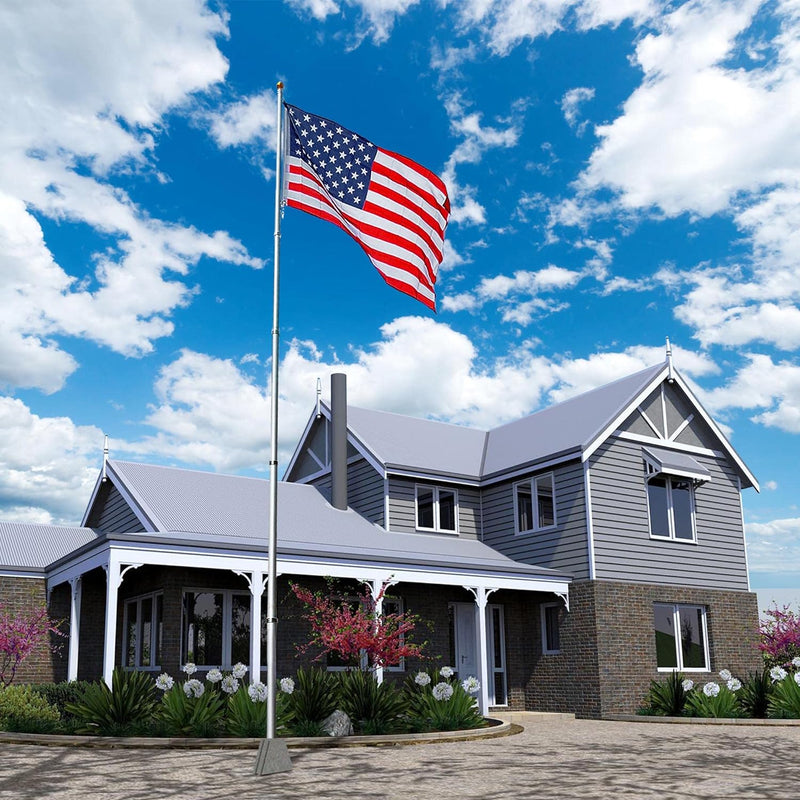 American flag proudly waves atop a pole in front of a modern gray house, surrounded by colorful flowers and a blue sky.