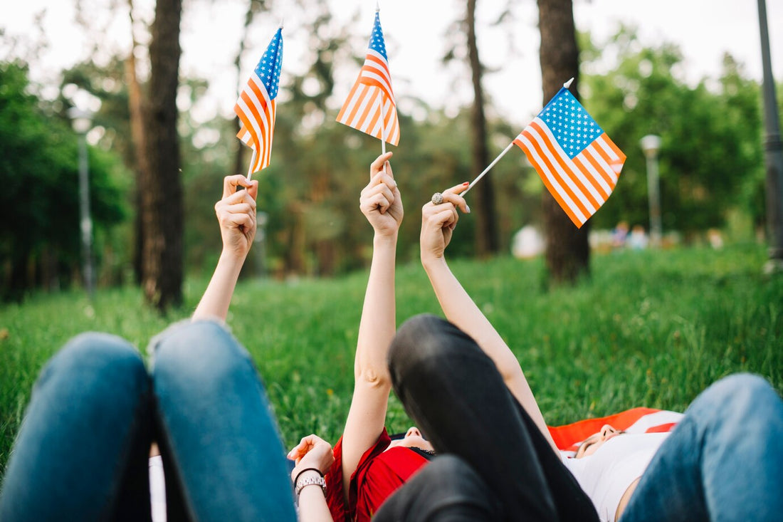 Three individuals lying on the grass, holding small American flags, celebrating patriotism in a lush park setting.